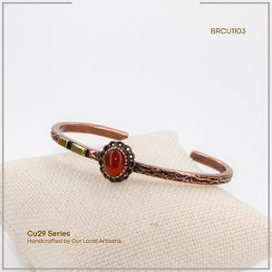Pu&ograve; includere: Bracciale a polsino in rame con una gemma ovale rosso-arancio. Il bracciale presenta un motivo floreale ed &egrave; impreziosito da quadrati oro e neri. Il testo "BRCU1103" &egrave; visibile. Il testo "Cu29 Series Handcrafted by Our Local Artisans" &egrave; anch'esso visibile.