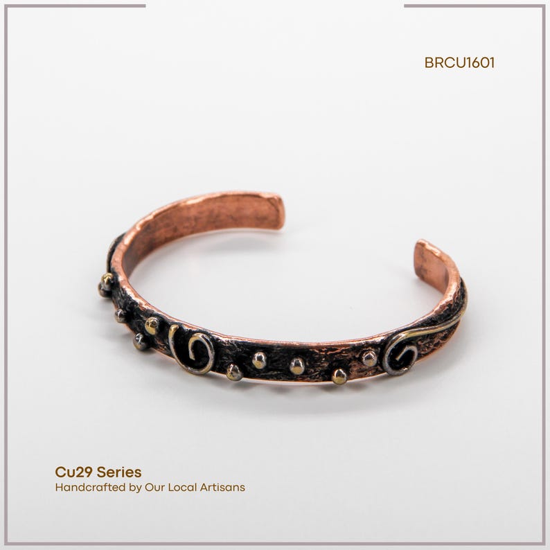 May include: Copper cuff bracelet with a dark, textured band and decorative swirls and small gold-coloured accents. The bracelet is part of the Cu29 Series and is handcrafted by local artisans. The text "BRCU1601" is also visible.