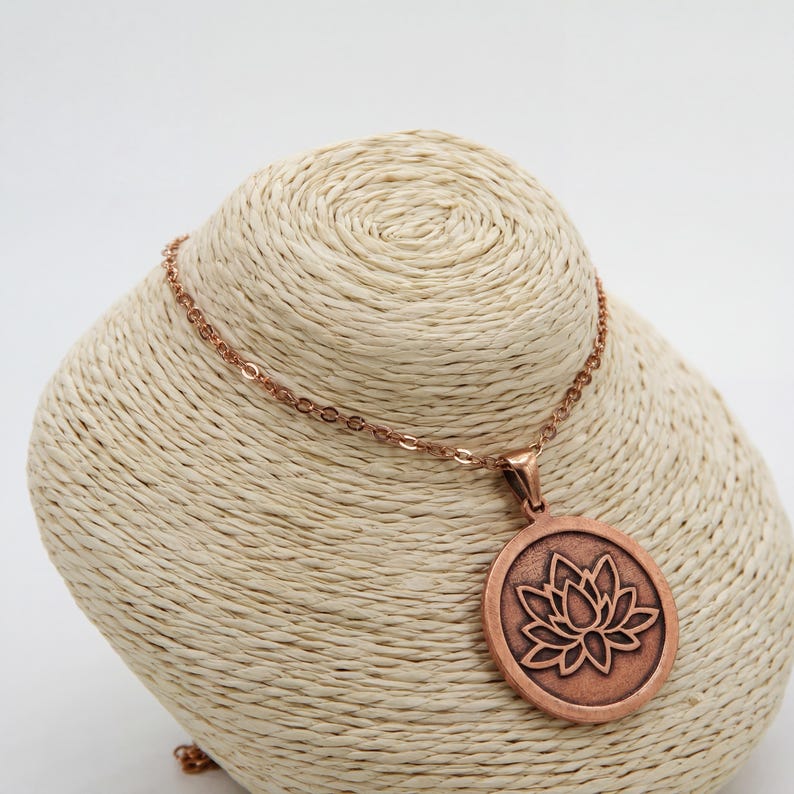 May include: Copper-colored necklace featuring a round pendant with a lotus flower design. The pendant is encircled by a raised border and hangs from a delicate chain. The necklace is displayed on a beige bust.