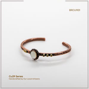 Pu&ograve; includere: Bracciale a polsino in rame con un elemento centrale in pietra ovale bianco sporco. Il bracciale presenta dettagli intricati e piccoli accenti dorati. Il testo "Cu29 Series" e "Handcrafted by Our Local Artisans" sono visibili.