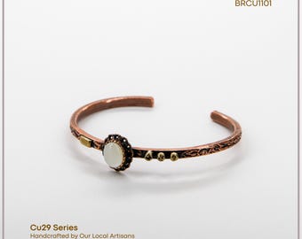 Può includere: Bracciale a polsino in rame con un elemento centrale in pietra ovale bianco sporco. Il bracciale presenta dettagli intricati e piccoli accenti dorati. Il testo "Cu29 Series" e "Handcrafted by Our Local Artisans" sono visibili.