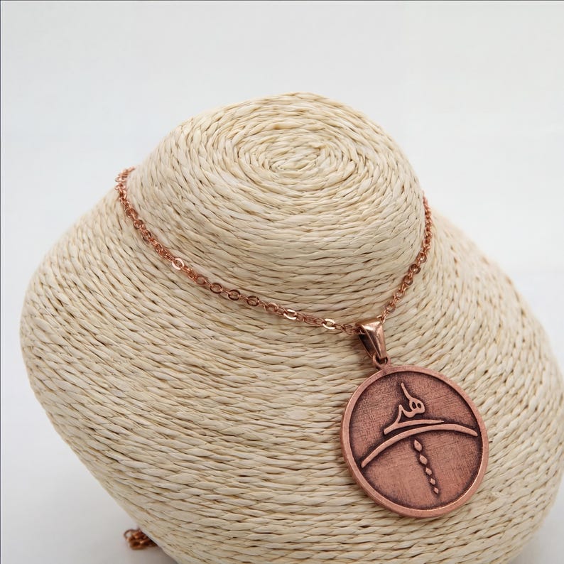 Op de afbeelding: Een koperkleurige hangerketting. De hanger heeft een cirkelvorm met een reli&euml;f ontwerp. De ketting en hanger hebben een bijpassende koperkleur. De ketting wordt gepresenteerd op een beige buste.