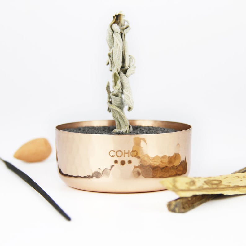 May include: A copper bowl with a hammered finish and the word "COHO" engraved on the side. The bowl is filled with black sand and a dried sage bundle is placed in the centre.
