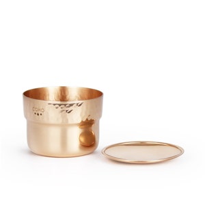May include: A hammered copper container with a lid. The container has a small hole with three dots and the word "COHO" inscribed on the side.
