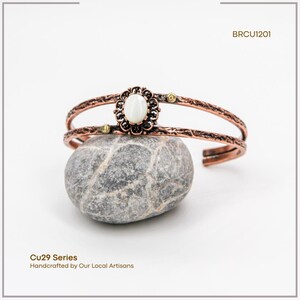 Puede incluir: Brazalete de pu&ntilde;o color cobre con una piedra preciosa ovalada blanca y detalles florales. El brazalete se muestra sobre una piedra gris. El texto "BRCU1201" es visible. El texto "Cu29 Series Handcrafted by Our Local Artisans" tambi&eacute;n es visible.