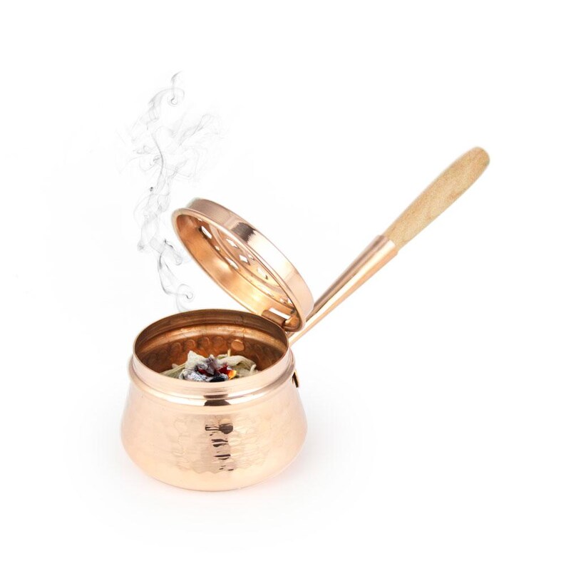 May include: A copper incense burner with a hammered finish and a wooden handle. The burner has a lid with a small hole in the center. Smoke is rising from the burner.