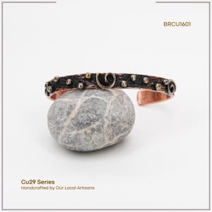 May include: A copper cuff bracelet with a dark, textured band, decorated with small gold and silver-toned studs and spiral designs. The bracelet is displayed on a grey stone, with the text "BRCU1601" visible. The text "Cu29 Series Handcrafted by Our Local Artisans" is also visible.