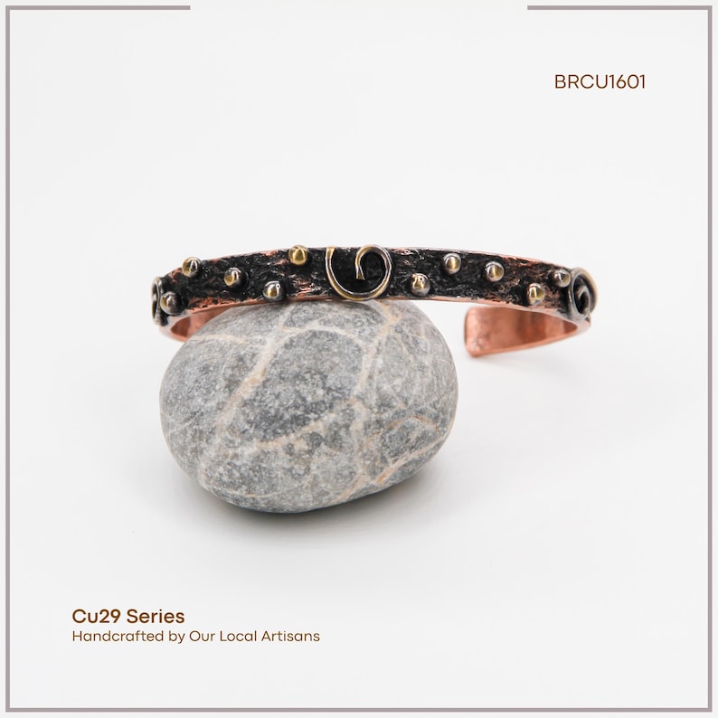 May include: A copper cuff bracelet with a dark, textured band, decorated with small gold and silver-toned studs and spiral designs. The bracelet is displayed on a grey stone, with the text "BRCU1601" visible. The text "Cu29 Series Handcrafted by Our Local Artisans" is also visible.
