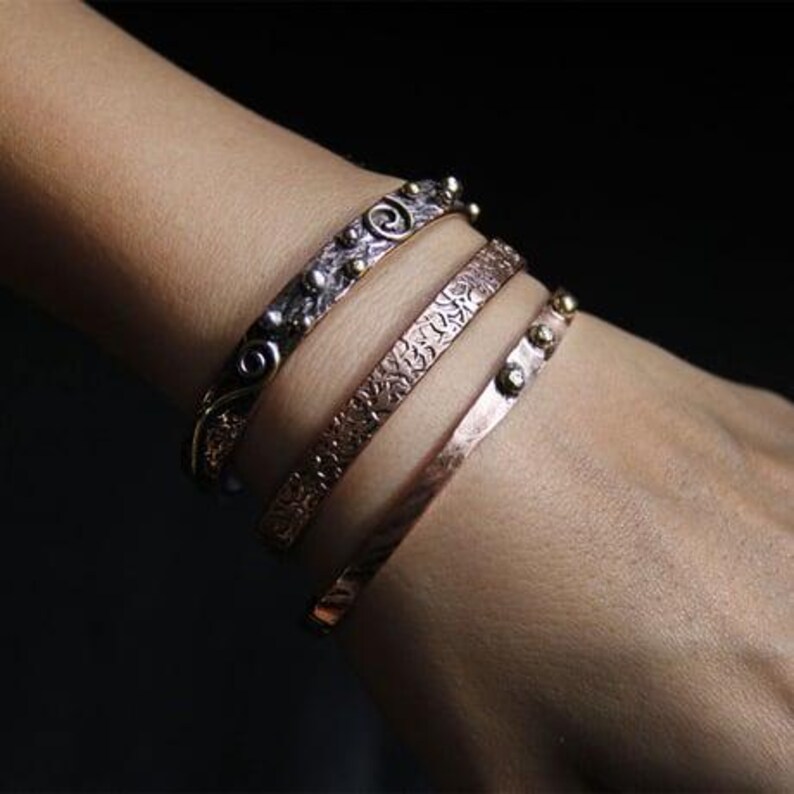 May include: Four metal bracelets in different designs and colours are stacked on a wrist. The bracelets include silver, copper, and bronze tones. One bracelet has a floral pattern, another has a swirl design, and others have small, round accents.