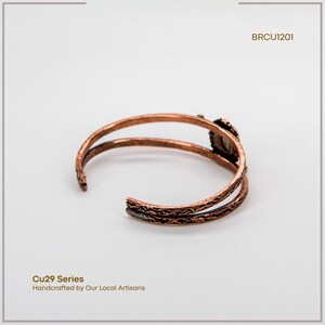 Puede incluir: Brazalete de pu&ntilde;o de cobre con un dise&ntilde;o abierto. El brazalete presenta un patr&oacute;n texturizado y un engaste de piedra decorativo. El texto "BRCU1201" es visible. El texto "Cu29 Series Handcrafted by Our Local Artisans" tambi&eacute;n es visible.