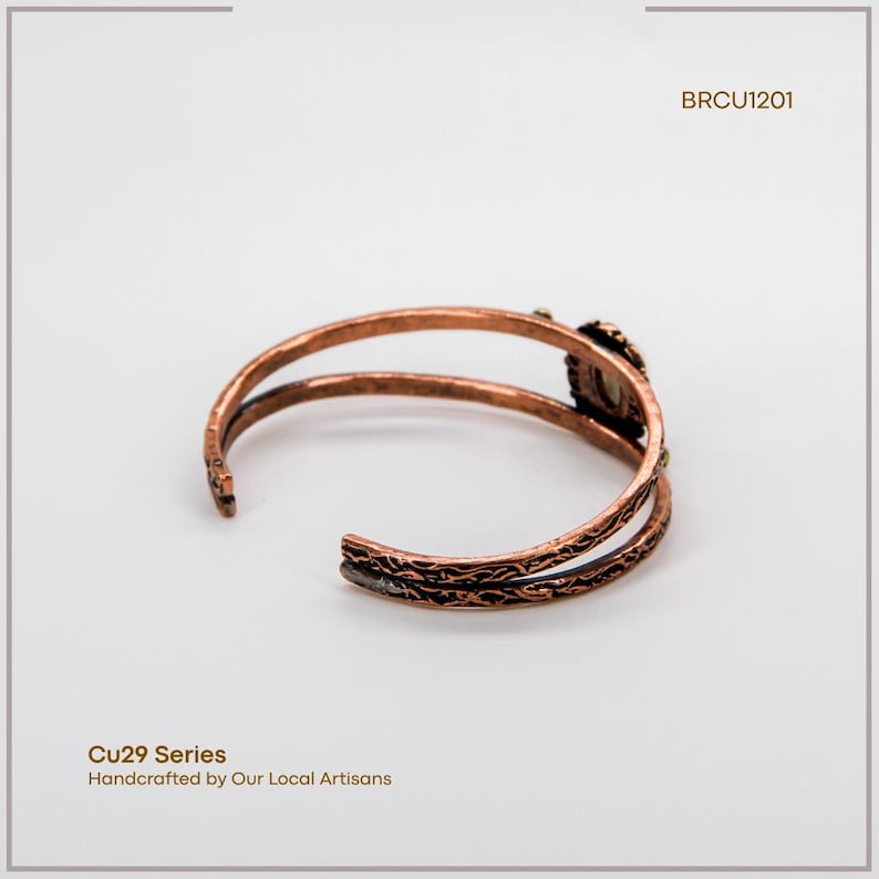 Puede incluir: Brazalete de pu&ntilde;o de cobre con un dise&ntilde;o abierto. El brazalete presenta un patr&oacute;n texturizado y un engaste de piedra decorativo. El texto "BRCU1201" es visible. El texto "Cu29 Series Handcrafted by Our Local Artisans" tambi&eacute;n es visible.