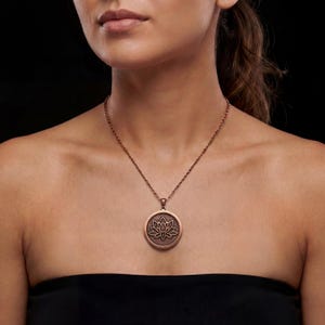 May include: A copper-colored necklace featuring a circular pendant with a lotus flower design. The pendant is suspended from a delicate chain. The necklace is worn against a black background.