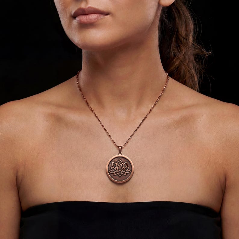 May include: A copper-colored necklace featuring a circular pendant with a lotus flower design. The pendant is suspended from a delicate chain. The necklace is worn against a black background.