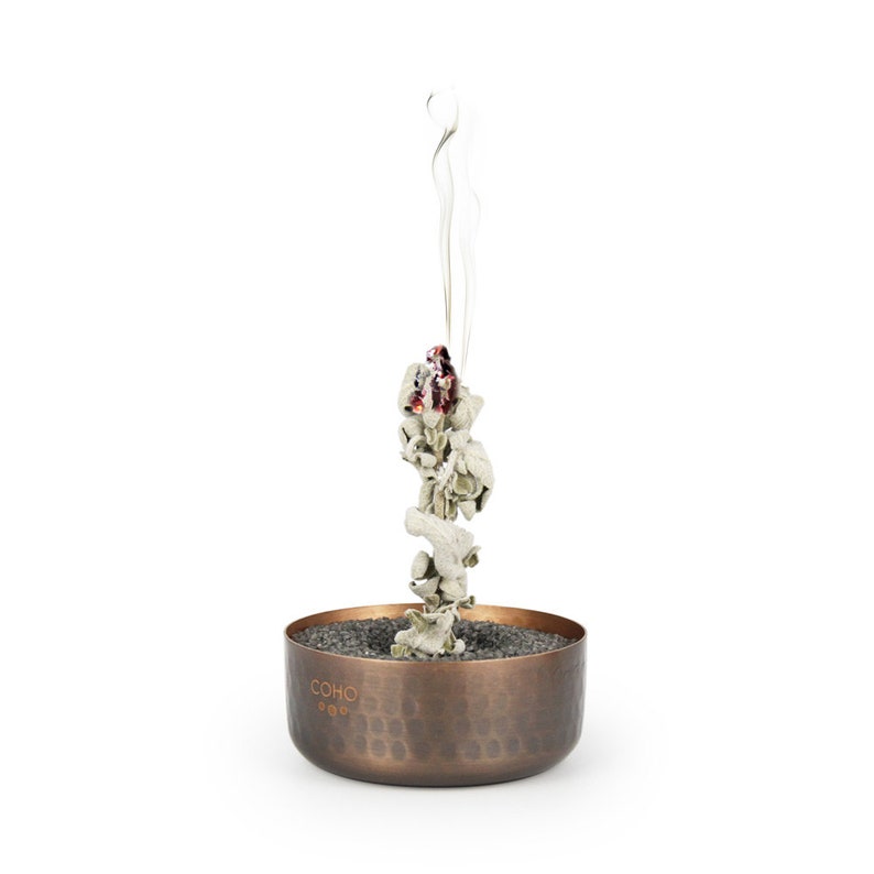 May include: A copper incense burner with a hammered finish and the word "COHO" engraved on the side. White incense is burning in the burner, producing a plume of smoke.