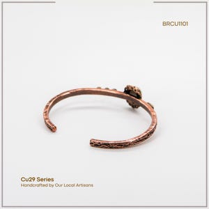 Pu&ograve; includere: Un bracciale a polsino in rame con superficie strutturata ed elemento decorativo. Il bracciale &egrave; aperto e presenta il testo "Cu29 Series Handcrafted by Our Local Artisans" e il codice "BRCU1101".