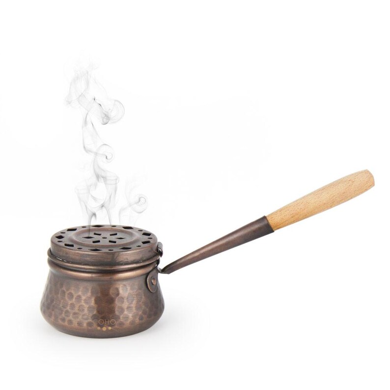 May include: A copper incense burner with a hammered finish and a wooden handle. Smoke rises from the burner.