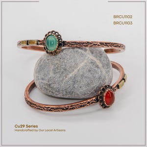 Pu&ograve; includere: Due braccialetti color rame con dettagli ornati e accenti di pietre preziose. Uno presenta una pietra verde, l'altro una pietra rossa. I braccialetti poggiano su una pietra grigia. Testo: "Cu29 Series Handcrafted by Our Local Artisans."