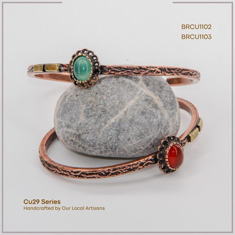 Pu&ograve; includere: Due braccialetti color rame con dettagli ornati e accenti di pietre preziose. Uno presenta una pietra verde, l'altro una pietra rossa. I braccialetti poggiano su una pietra grigia. Testo: "Cu29 Series Handcrafted by Our Local Artisans."