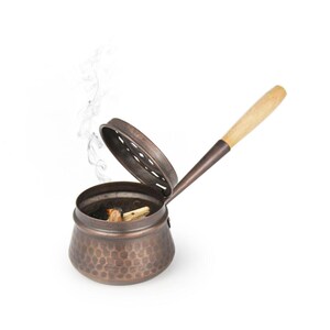 May include: A copper incense burner with a wooden handle and a lid with holes. The burner is filled with incense sticks and smoke is rising from the top.