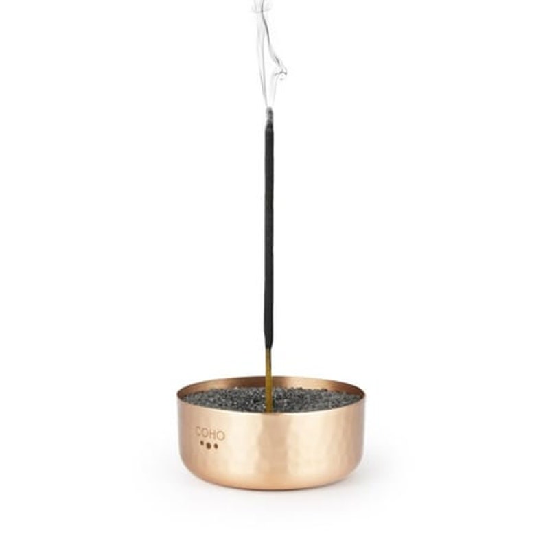 May include: A copper incense holder with a hammered finish and three small holes. A black incense stick is burning in the holder, with smoke rising from the top.