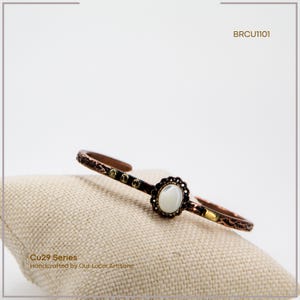 Pu&ograve; includere: Bracciale a polsino color rame con una pietra centrale ovale bianco sporco e un design traforato decorativo. Fa parte della serie Cu29, realizzato a mano da artigiani locali. &Egrave; visibile anche il testo "BRCU1101".