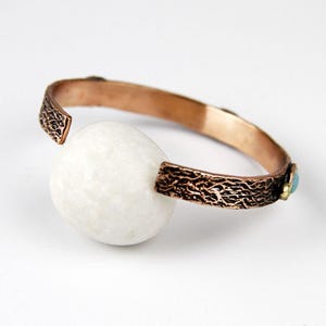 May include: A copper bracelet with a textured finish and a white stone accent. The bracelet is decorated with small turquoise and gold accents.