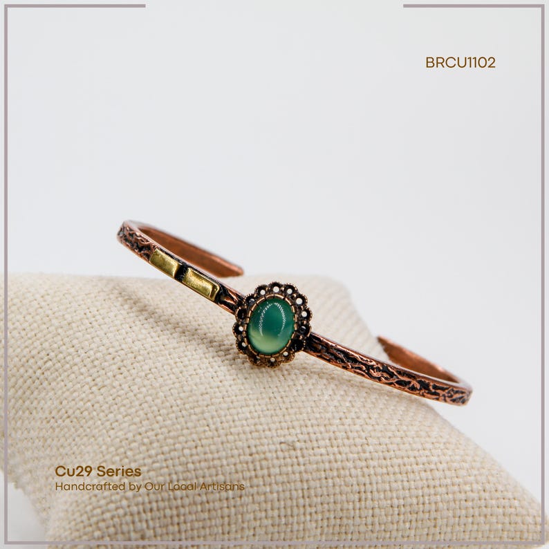 Pu&ograve; includere: Bracciale a polsino in rame con una pietra verde ovale al centro, circondata da un motivo floreale decorativo. Il bracciale presenta un motivo strutturato e il testo "BRCU1102" e "Cu29 Series Handcrafted by Our Local Artisans."