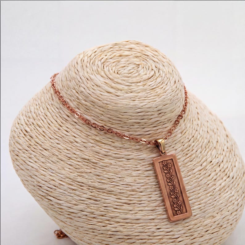 Puede incluir: Un collar de tono cobre con un colgante rectangular que presenta un patr&oacute;n de enredadera floral en relieve. El colgante cuelga de una delicada cadena a juego, mostrando un acabado met&aacute;lico y un dise&ntilde;o vertical con flores.