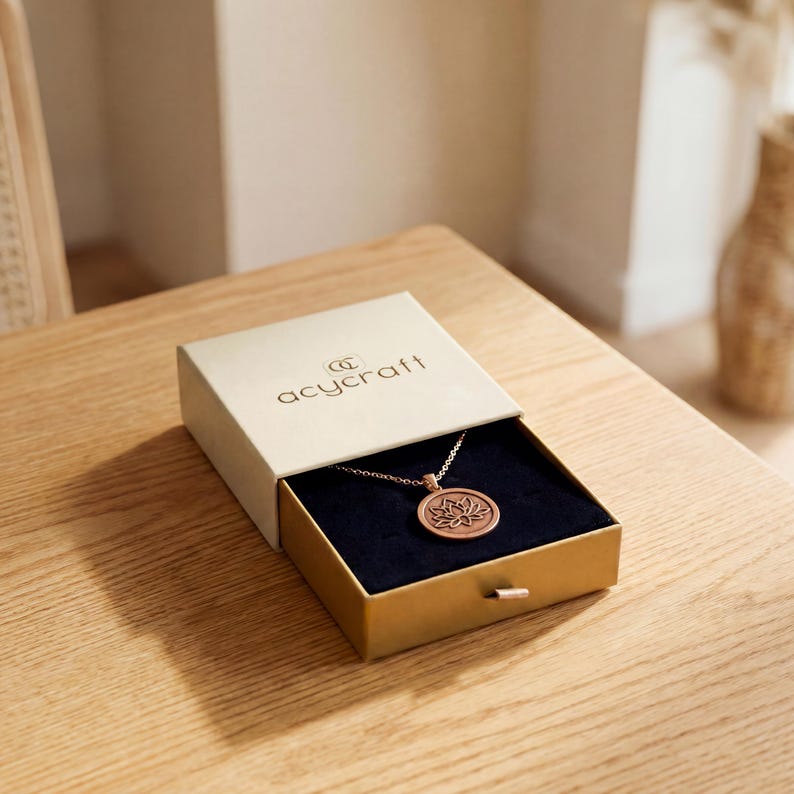 May include: A gold-colored necklace with a lotus flower pendant rests inside an open gift box. The box is beige with the brand name "acycraft" printed on the lid. The box is on a wooden surface.