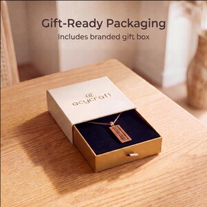 Puede incluir: Un collar con colgante rectangular de acabado met&aacute;lico descansa dentro de una caja de regalo deslizante de color beige. La tapa muestra el texto Gift-Ready Packaging, Includes branded gift box y la marca acycraft en marr&oacute;n.