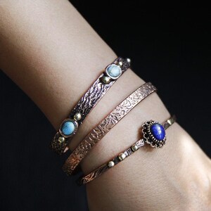 May include: Three copper bracelets with textured surfaces and gemstone accents. The top bracelet has two turquoise stones, the middle bracelet has a turquoise stone, and the bottom bracelet has a lapis lazuli stone.