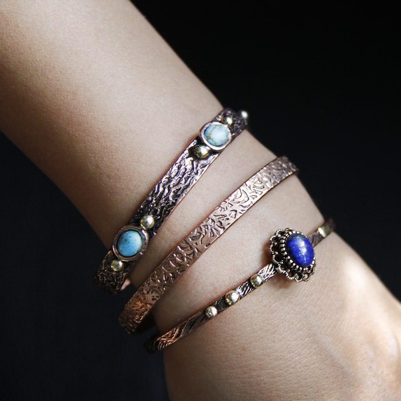 May include: Three copper bracelets with textured surfaces and gemstone accents. The top bracelet has two turquoise stones, the middle bracelet has a turquoise stone, and the bottom bracelet has a lapis lazuli stone.