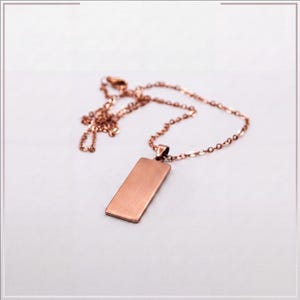 Puede incluir: Un collar de tono oro rosa con un colgante rectangular de acabado met&aacute;lico cepillado. El colgante est&aacute; unido a una delicada cadena a juego del mismo tono, formada por peque&ntilde;os eslabones ovalados uniformes de dise&ntilde;o sencillo y limpio.