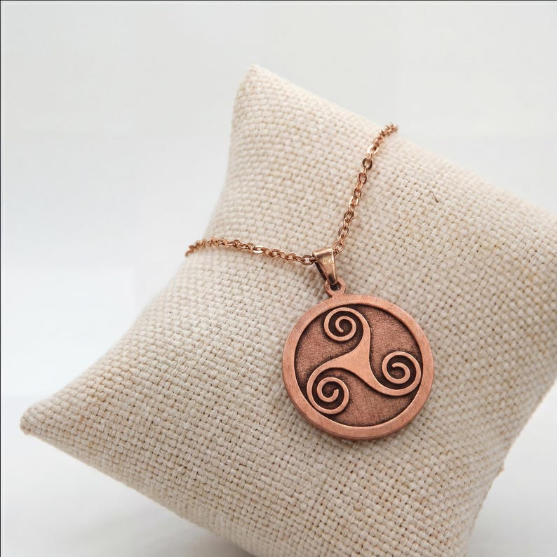 May include: A copper-coloured pendant necklace featuring a circular pendant with a Celtic Triskele design. The pendant hangs from a delicate chain. The necklace is displayed on a beige, textured cushion.