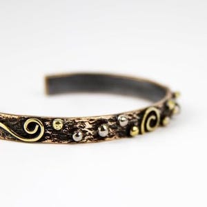 May include: A bronze cuff bracelet with a textured surface and decorative accents. The bracelet features gold swirls and small silver and gold beads. The interior of the bracelet is dark grey.