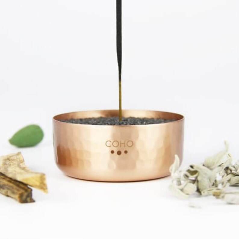 May include: A copper incense holder with a hammered finish. The holder has a black incense stick inserted into it. The holder has the word "COHO" engraved on it.