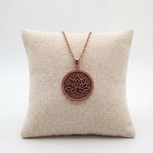 May include: A copper-colored necklace featuring a circular pendant with a lotus flower design. The pendant hangs from a delicate chain. The necklace is displayed on a beige, textured pillow against a white background.