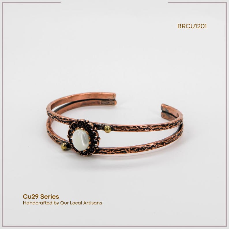 Puede incluir: Brazalete de pu&ntilde;o de cobre con un dise&ntilde;o floral y una piedra blanca ovalada en el centro. El brazalete tiene dos bandas paralelas y peque&ntilde;os detalles dorados. El texto "Cu29 Series" y "Handcrafted by Our Local Artisans" es visible.