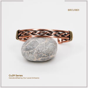 May include: Copper cuff bracelet with a braided design, resting on a gray stone. The bracelet has a warm, reddish-brown hue with gold-colored accents on the ends. The text "BRCU1801" is visible in the upper right corner.