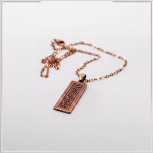 Puede incluir: Un collar con colgante rectangular de tono cobre que presenta un patr&oacute;n de enredadera floral en relieve. El colgante cuelga de una cadena de metal a juego de color cobre con peque&ntilde;os eslabones ovalados. El dise&ntilde;o muestra flores.