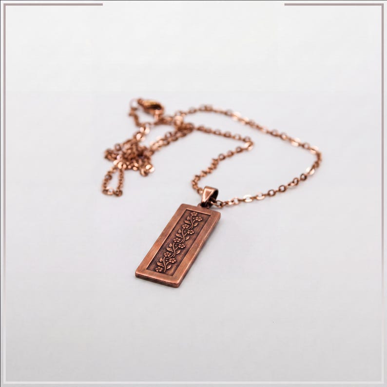 Puede incluir: Un collar con colgante rectangular de tono cobre que presenta un patr&oacute;n de enredadera floral en relieve. El colgante cuelga de una cadena de metal a juego de color cobre con peque&ntilde;os eslabones ovalados. El dise&ntilde;o muestra flores.