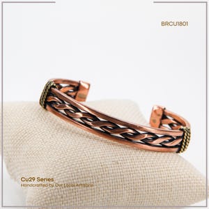 May include: Copper-colored bracelet with a braided design, resting on a beige surface. The bracelet has a woven pattern and is accented with gold-colored details. The text "Cu29 Series Handcrafted by Our Local Artisans" is visible.