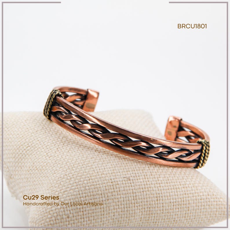 May include: Copper-colored bracelet with a braided design, resting on a beige surface. The bracelet has a woven pattern and is accented with gold-colored details. The text "Cu29 Series Handcrafted by Our Local Artisans" is visible.
