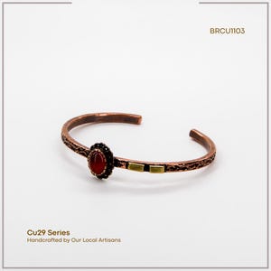 Pu&ograve; includere: Bracciale a polsino in rame con una gemma rossa ovale e dettagli decorativi. Il bracciale presenta un design strutturato e due piccoli accenti rettangolari color oro. Il testo "Cu29 Series Handcrafted by Our Local Artisans" &egrave; visibile.
