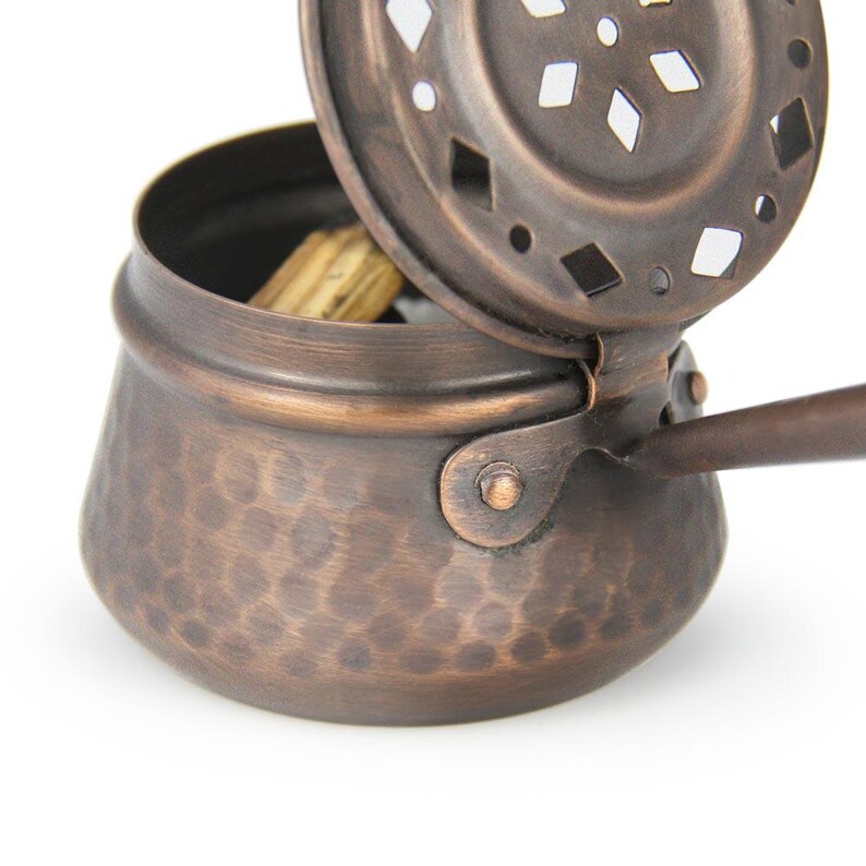 May include: A copper incense burner with a hammered finish and a lid with a decorative cut-out design. The burner has a handle and is open, revealing incense sticks inside.