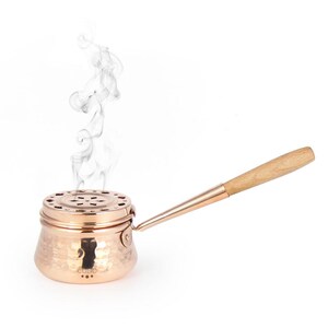 May include: A copper incense burner with a hammered finish and a wooden handle. Smoke rises from the burner.