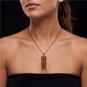 Puede incluir: Un collar con colgante rectangular en tono cobre que presenta un patr&oacute;n floral en relieve. El colgante cuelga de una fina cadena met&aacute;lica a juego, mostrando un dise&ntilde;o vertical con detalles florales intrincados sobre un fondo oscuro.