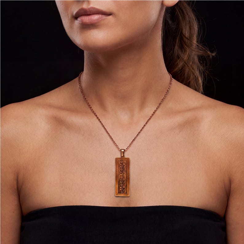 Puede incluir: Un collar con colgante rectangular en tono cobre que presenta un patr&oacute;n floral en relieve. El colgante cuelga de una fina cadena met&aacute;lica a juego, mostrando un dise&ntilde;o vertical con detalles florales intrincados sobre un fondo oscuro.
