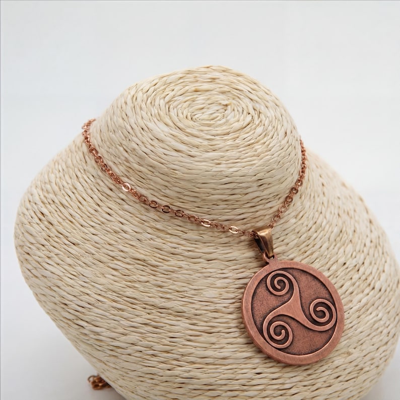 May include: A copper-toned pendant necklace with a circular pendant displaying a Celtic triskele design. The necklace is presented on a mannequin bust. The chain and pendant share a metallic finish, creating a coordinated and fashionable accessory.