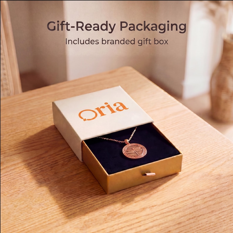 Pode incluir: Um colar de ouro rosa com um pingente circular de lib&eacute;lula dentro de uma caixa de presente de marca branca e bege entreaberta. A tampa apresenta o texto Oria em laranja, com Gift-Ready Packaging Includes branded gift box escrito acima.